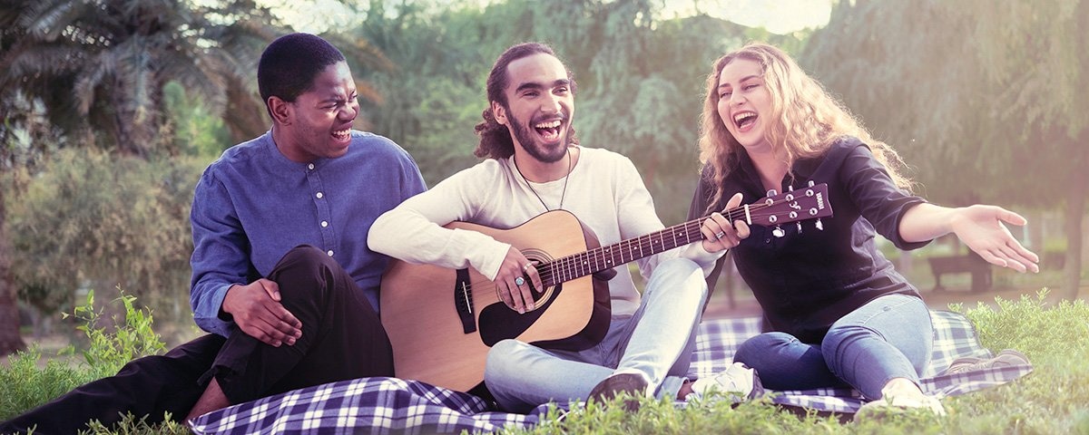 Three smiling people sitting on a blanket outdoors. Middle person is playing an F310 acoustic guitar.
